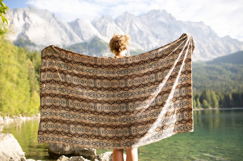 May include: A large, rectangular blanket with a snakeskin pattern in shades of brown and tan is held up against a backdrop of mountains, a lake, and trees. The blanket is draped, showcasing its design and texture.
