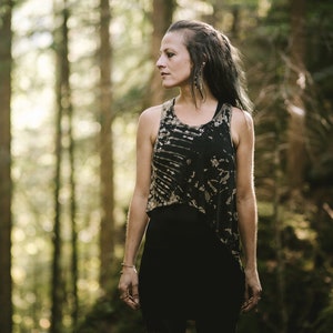 Puede incluir: Una mujer con el pelo largo y oscuro se encuentra en un bosque, vistiendo un top sin mangas de color negro y marrón con efecto tie-dye y pantalones negros.