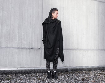 May include: A person wearing a long, black hooded cardigan with a draped design. The cardigan is worn over a pair of black and white patterned leggings. The person is standing in front of a gray concrete wall.
