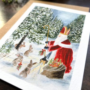May include: A greeting card featuring a watercolor illustration of a figure in a red robe and hat, resembling Santa Claus, in a snowy forest. Rabbits gather around him. The card is on a brown envelope.