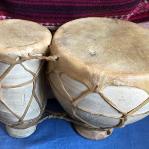 May include: A pair of handmade bongo drums. The drums have light tan leather drumheads stretched over white ceramic bodies. The drumheads are secured with a crisscross pattern of tan rope. The drums are set against a blue surface.