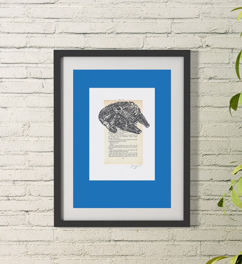 K&ouml;nnte beinhalten: Eine Schwarzwei&szlig;illustration des Raumschiffs Millennium Falcon aus Star Wars, gedruckt auf einer Buchseite. Das Bild ist in einem schwarzen Rahmen mit einer blauen Passepartout gerahmt.