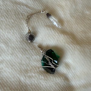 May include: A decorative hanging ornament featuring a large, raw, dark green stone wrapped in silver-toned wire. Attached to the wire is a silver-toned metal chain holding a small, dark purple spherical bead in a wire cage and a clear, pointed crystal.