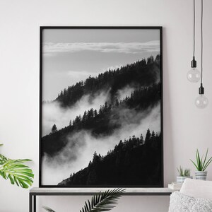 May include: Black and white photograph of a mountain range with fog rolling through the trees. The photo is framed in a black frame and hanging on a white wall.