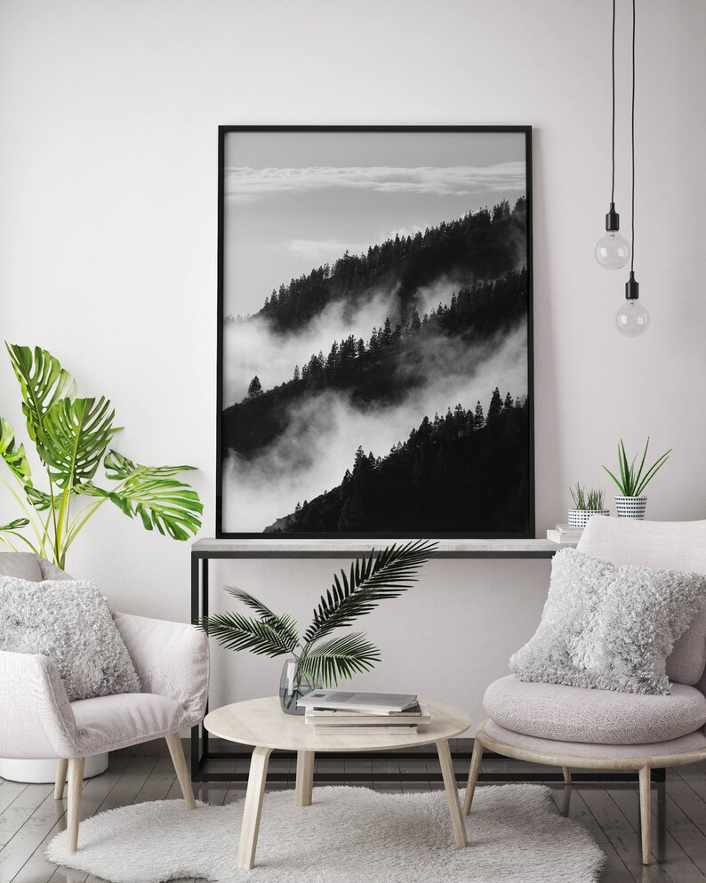 May include: Black and white photograph of a mountain range with fog rolling through the trees. The photo is framed in a black frame and hanging on a white wall.