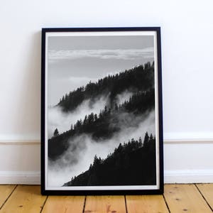 May include: Black and white photograph of a mountain range with fog rolling through the trees.
