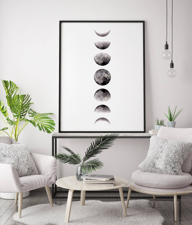 May include: A black and white print of the moon phases in various stages, framed in a black frame and hanging on a white wall above a white couch and a coffee table with a plant on it.