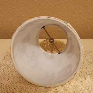 May include: A cylindrical lamp shade featuring a white background with a repeating pattern of dark blue silhouettes depicting figures and horse-drawn carriages. The interior includes a gold-toned metal wire spider fitter assembly for mounting.