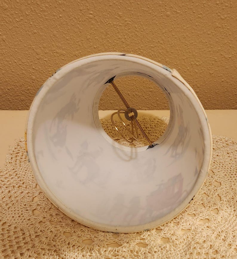 May include: A cylindrical lamp shade featuring a white background with a repeating pattern of dark blue silhouettes depicting figures and horse-drawn carriages. The interior includes a gold-toned metal wire spider fitter assembly for mounting.