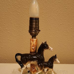 May include: A table lamp featuring a ceramic base with a mare and foal standing on a green grassy patch. The central post resembles a tree trunk, topped with a brass-colored socket and a single light bulb. The horses are black with white markings.