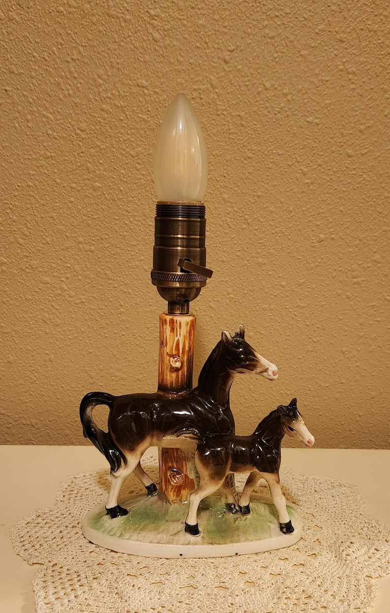 May include: A table lamp featuring a ceramic base with a mare and foal standing on a green grassy patch. The central post resembles a tree trunk, topped with a brass-colored socket and a single light bulb. The horses are black with white markings.