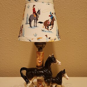 May include: A table lamp featuring a ceramic base with a black and white mare and foal standing on a green grassy oval platform. The central post resembles a tree trunk. The fabric lampshade displays a colorful pattern of cowboys on horseback.