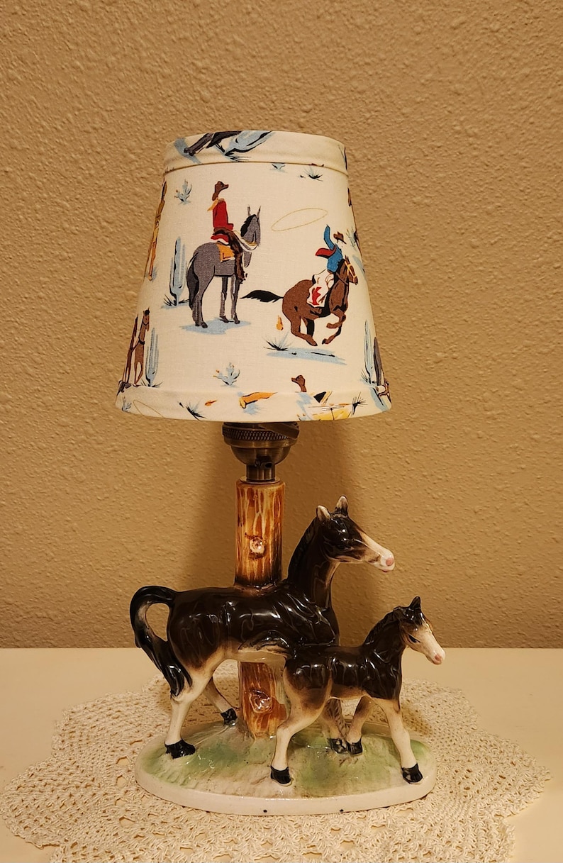 May include: A table lamp featuring a ceramic base with a black and white mare and foal standing on a green grassy oval platform. The central post resembles a tree trunk. The fabric lampshade displays a colorful pattern of cowboys on horseback.