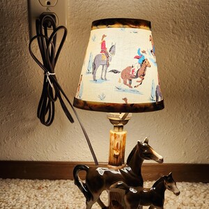 May include: A table lamp featuring a ceramic base with a brown horse and foal standing next to a tree trunk. The lamp has a conical shade with a cream background and a colorful western-themed print of riders on horseback and cacti.