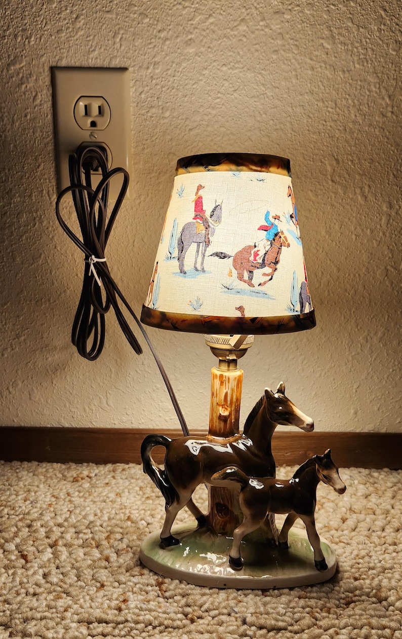 May include: A table lamp featuring a ceramic base with a brown horse and foal standing next to a tree trunk. The lamp has a conical shade with a cream background and a colorful western-themed print of riders on horseback and cacti.