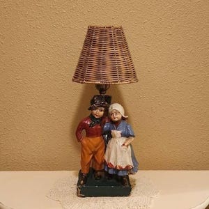 May include: A vintage lamp featuring a woven lampshade and a base with two figurines dressed in traditional attire. The figurines are a boy in orange pants and a red shirt, and a girl in a blue dress and white apron.