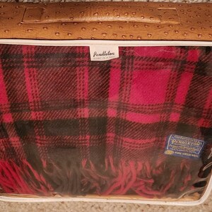 May include: A Pendleton wool blanket with a red and black plaid pattern, featuring fringe edges. The blanket is folded inside a rectangular, tan-colored storage case with a textured, ostrich-skin-like finish and a clear plastic front window.