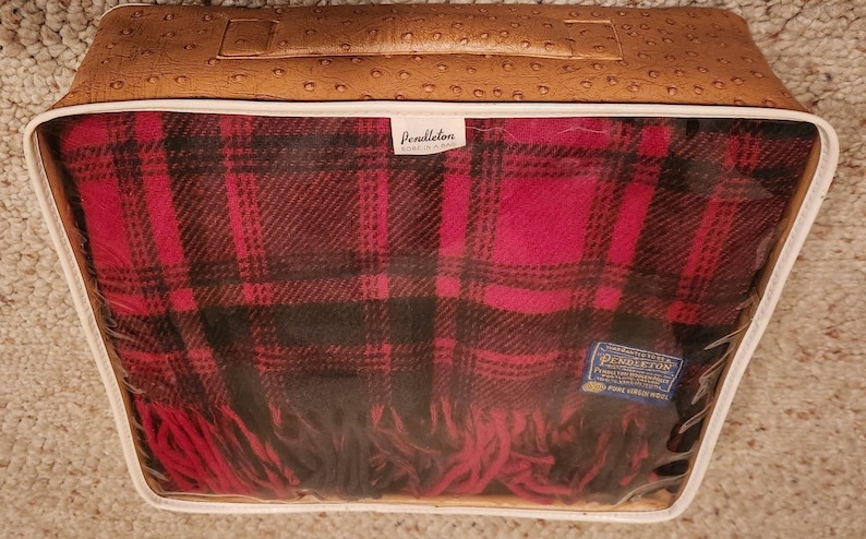 May include: A Pendleton wool blanket with a red and black plaid pattern, featuring fringe edges. The blanket is folded inside a rectangular, tan-colored storage case with a textured, ostrich-skin-like finish and a clear plastic front window.