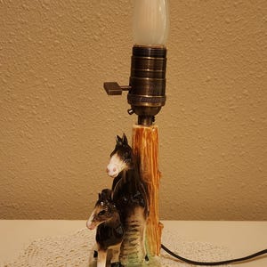 May include: A ceramic table lamp base featuring a sculpted brown and white horse and foal standing beside a textured tree trunk. The lamp has a metallic bronze-toned socket with a turn-knob switch and a single white flame-shaped light bulb.