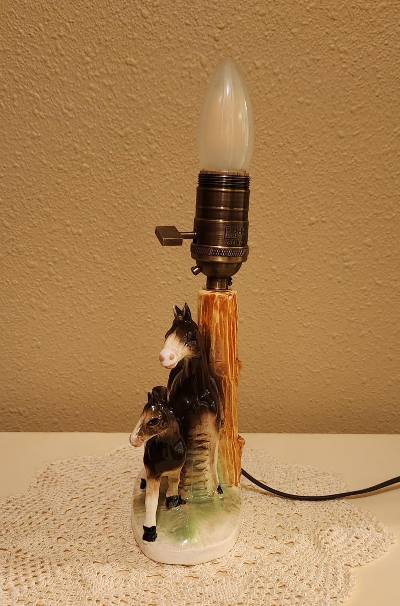 May include: A ceramic table lamp base featuring a sculpted brown and white horse and foal standing beside a textured tree trunk. The lamp has a metallic bronze-toned socket with a turn-knob switch and a single white flame-shaped light bulb.