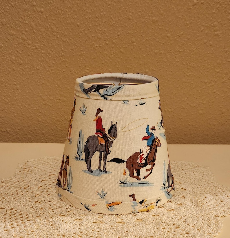 May include: A tapered lamp shade with a cream-colored background featuring a repeating pattern of cowboys riding horses, cacti, and lassos in shades of red, blue, brown, and grey. The shade has a metal fitter ring at the top and finished edges.