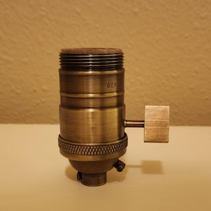 May include: A vintage brass lamp socket with a wooden top and a square switch. The socket has a textured design and the text "02AL" is visible. This lighting component is designed for use in lamps.