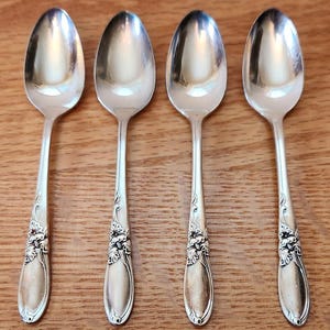 May include: Four silver spoons with oval bowls and ornate floral designs on the handles. The spoons are arranged on a wooden surface, showcasing their reflective surfaces and intricate detailing. A classic silverware set.