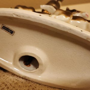 May include: An oval-shaped ceramic base for a lamp featuring a white finish with a small rectangular sticker that reads JAPAN. The base has a central circular opening for a cord and displays a partial view of brown and white animal figurines.