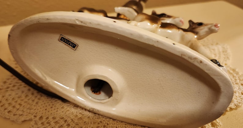 May include: An oval-shaped ceramic base for a lamp featuring a white finish with a small rectangular sticker that reads JAPAN. The base has a central circular opening for a cord and displays a partial view of brown and white animal figurines.