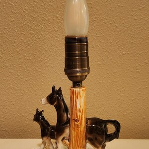May include: A ceramic table lamp featuring a dark brown and white donkey and foal standing on a green base next to a textured, light brown tree trunk pillar. The lamp has a metallic bronze-toned socket and a single, frosted, flame-shaped light bulb.