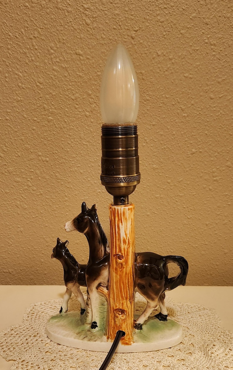 May include: A ceramic table lamp featuring a dark brown and white donkey and foal standing on a green base next to a textured, light brown tree trunk pillar. The lamp has a metallic bronze-toned socket and a single, frosted, flame-shaped light bulb.