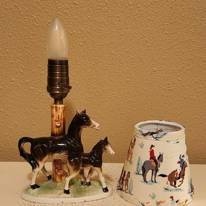 May include: A ceramic table lamp base featuring a dark brown mare and foal standing on a green grassy oval platform next to a faux-wood post. Beside the base sits a tapered fabric lampshade decorated with a colorful pattern of cowboys on horses.