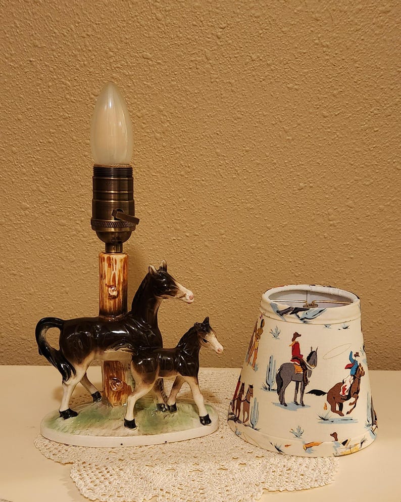 May include: A ceramic table lamp base featuring a dark brown mare and foal standing on a green grassy oval platform next to a faux-wood post. Beside the base sits a tapered fabric lampshade decorated with a colorful pattern of cowboys on horses.