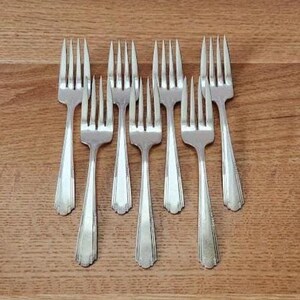 May include: A collection of eight silver forks arranged on a wooden surface. The forks have a classic design with tines and decorative handles. The arrangement suggests a formal dining setting or a vintage silverware set.