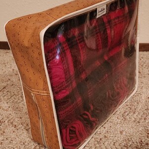 May include: A rectangular storage bag with a tan, ostrich-textured exterior and a clear plastic front panel. Inside, a folded red and black plaid blanket is visible. A white label with the text Pendleton is attached to the top interior edge.