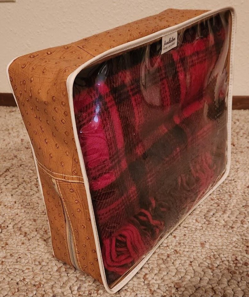 May include: A rectangular storage bag with a tan, ostrich-textured exterior and a clear plastic front panel. Inside, a folded red and black plaid blanket is visible. A white label with the text Pendleton is attached to the top interior edge.