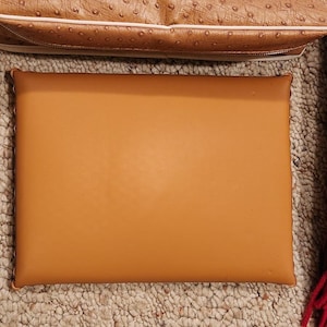 May include: A rectangular, smooth, tan-colored flat object with rounded corners and stitched edges. Positioned above it is a portion of a tan-colored bag with a textured, dotted surface and a white trim detail along the bottom edge.