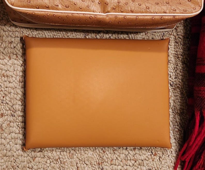 May include: A rectangular, smooth, tan-colored flat object with rounded corners and stitched edges. Positioned above it is a portion of a tan-colored bag with a textured, dotted surface and a white trim detail along the bottom edge.