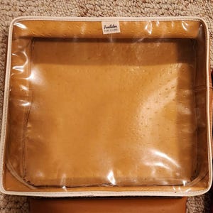 May include: A square storage bag with a tan, textured faux-ostrich leather finish and a clear plastic top panel. The bag features white piping along the edges and a small white label at the top center that reads Pendleton ROBE IN A BAG.