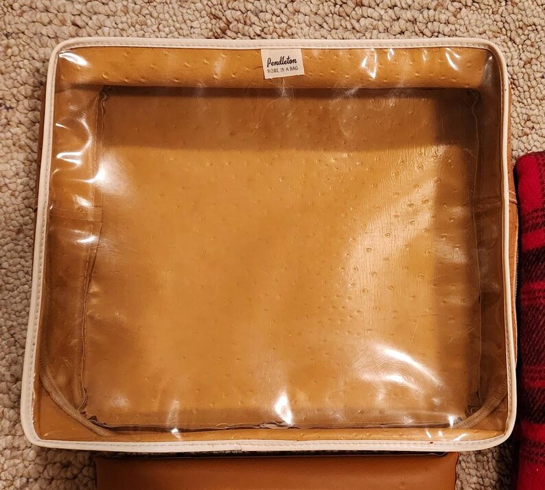 May include: A square storage bag with a tan, textured faux-ostrich leather finish and a clear plastic top panel. The bag features white piping along the edges and a small white label at the top center that reads Pendleton ROBE IN A BAG.
