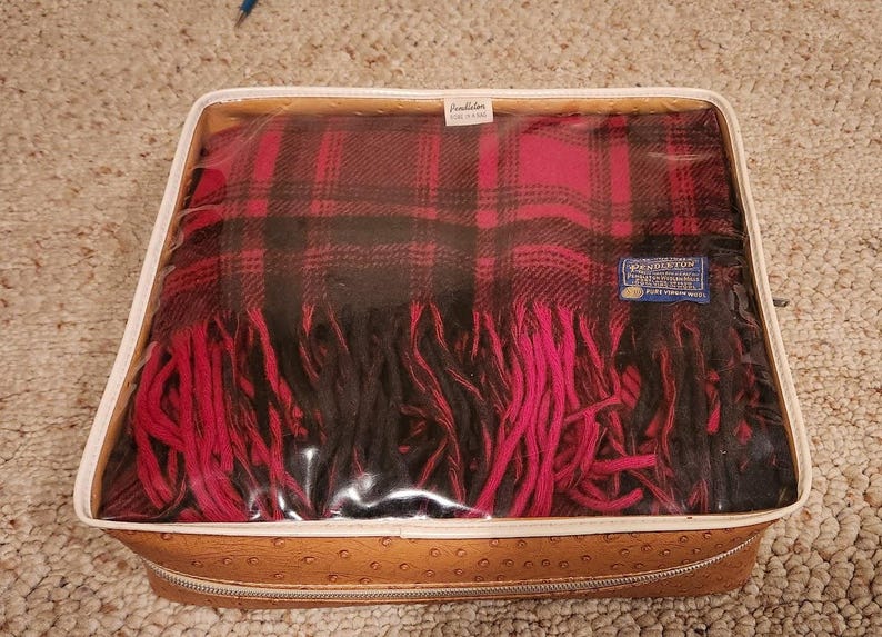 May include: A Pendleton plaid blanket in red and black, featuring a fringed edge and a blue label reading Pure Virgin Wool. The blanket is stored inside a rectangular, tan-colored, textured storage case with a clear plastic top and a zipper closure.