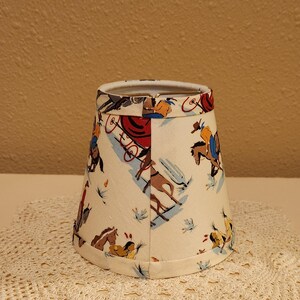 May include: A conical lamp shade with a cream-colored background featuring a repeating pattern of cowboys on horseback, stagecoaches, and figures in western attire. The fabric shade has a metal wire frame at the top and a finished hem at the bottom.