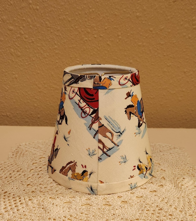 May include: A conical lamp shade with a cream-colored background featuring a repeating pattern of cowboys on horseback, stagecoaches, and figures in western attire. The fabric shade has a metal wire frame at the top and a finished hem at the bottom.