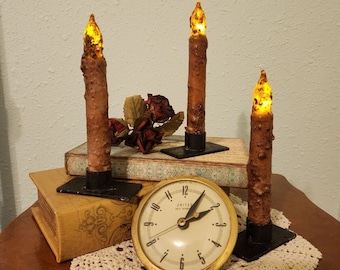 May include: Three rustic candle holders with lit candles on a wooden table with a clock and a doily. The candles are brown and have a yellow flame. The clock is gold and has black hands. The doily is white and lace.