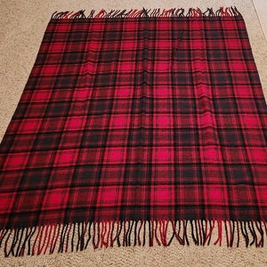 May include: A rectangular throw blanket featuring a red and black plaid tartan pattern. The blanket has a woven texture and is finished with long, fringed edges in alternating red and black yarn along the top and bottom sides of the fabric.