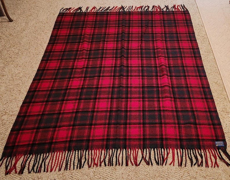 May include: A rectangular throw blanket featuring a red and black plaid tartan pattern. The blanket has a woven texture and is finished with long, fringed edges in alternating red and black yarn along the top and bottom sides of the fabric.