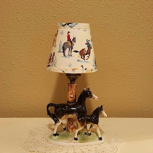 May include: A table lamp featuring a ceramic base with a dark brown mare and foal standing on a green grassy oval platform. The lamp post resembles a tree trunk. The conical lampshade is off-white with a colorful pattern of cowboys on horses.