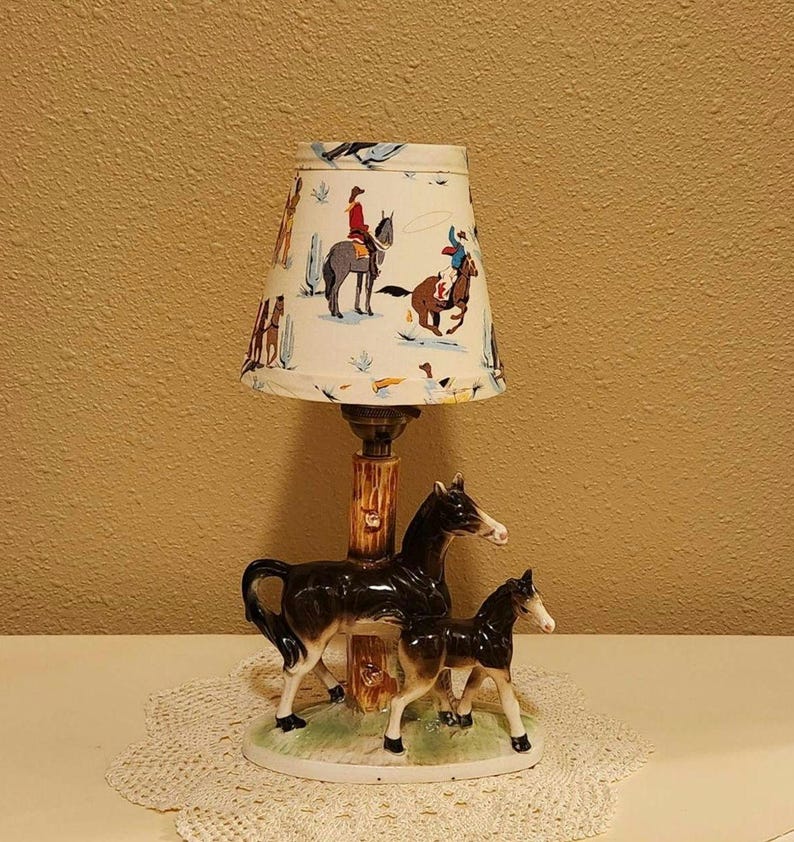 May include: A table lamp featuring a ceramic base with a dark brown mare and foal standing on a green grassy oval platform. The lamp post resembles a tree trunk. The conical lampshade is off-white with a colorful pattern of cowboys on horses.