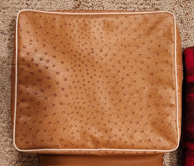 May include: A square-shaped tan bag featuring a textured ostrich-style pattern across the surface. The bag is outlined with a thin, light-colored piping along the edges, creating a defined border around the main tan panel of the accessory.