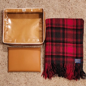 May include: A tan rectangular storage case with a clear plastic top, a solid tan rectangular pouch, and a folded red and black plaid wool blanket with fringe and a Pendleton brand label in the corner. The items are arranged on a textured surface.